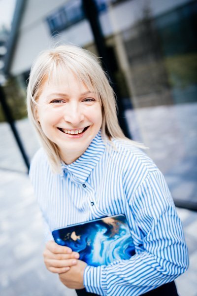 Nika Mak performance mindset coach smiling in a blue and white stripe shirt outside with laptop